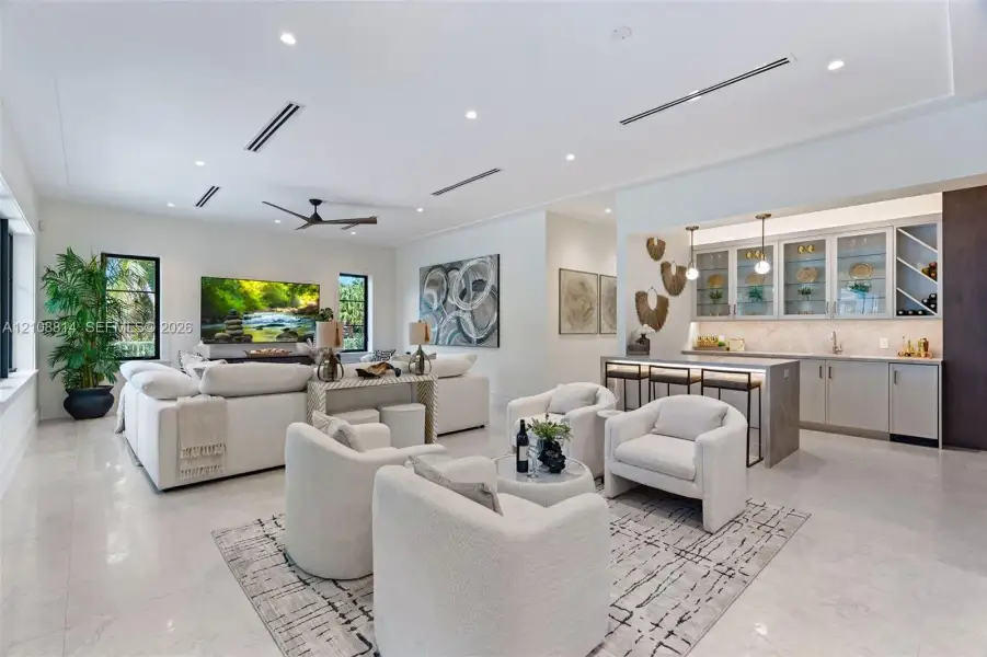 Open-concept living space featuring high ceilings with recessed lighting, large-format tile flooring, a dedicated wet bar with glass-front cabinetry, and a central island with integrated stone surfacing Open-concept living space featuring high ceilings with recessed lighting, large-format tile flooring, a dedicated wet bar with glass-front cabinetry, and a central island with integrated stone surfacing