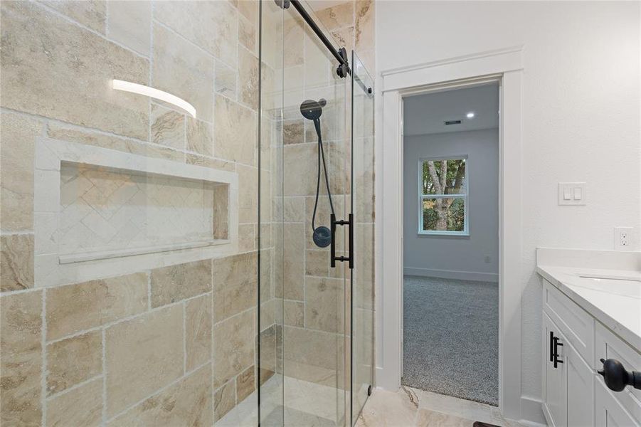 This is a modern bathroom featuring a spacious walk-in shower with stone tile walls and a glass door, complemented by a sleek built-in shelf. There is also a glimpse into a room with carpeting and a view of the outdoors through a window.