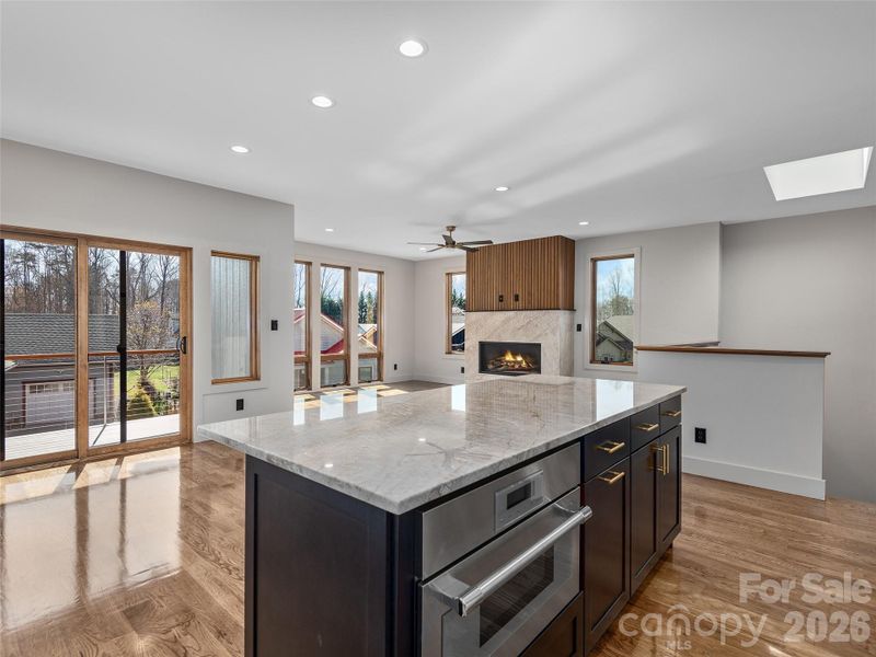 View from the kitchen across the open-concept main living area, highlighting the oversized island, oak hardwood floors, vented gas fireplace, and sliding doors to the covered porch.