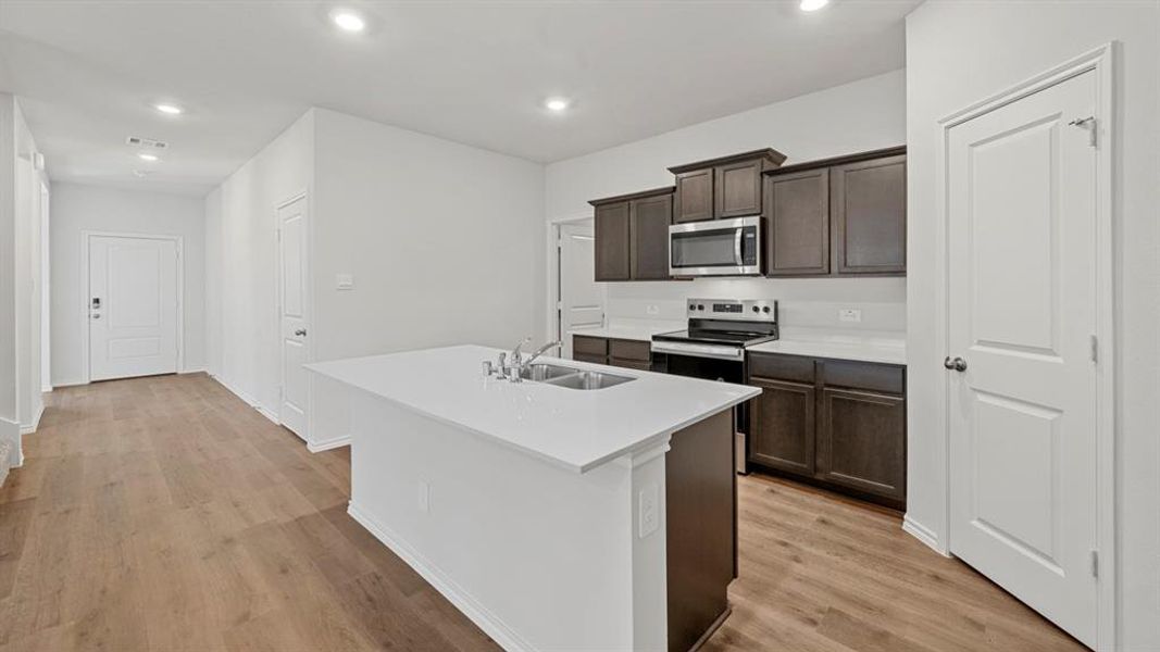 Kitchen with dark brown cabinets, stainless steel appliances, light wood-style flooring, a center island with sink, and recessed lighting
