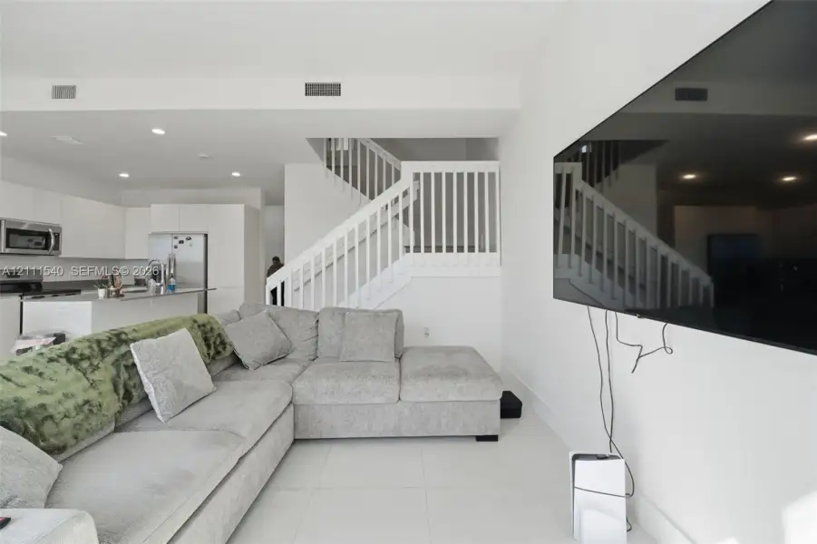 Open-concept interior featuring light-toned tile flooring, recessed ceiling lighting, a staircase with white balusters, and a kitchen equipped with white cabinetry