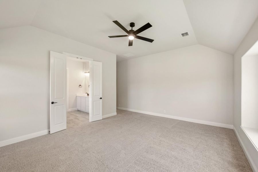 Empty bedroom with vaulted ceiling, ceiling fan, carpet flooring, and open door to ensuite bathroom