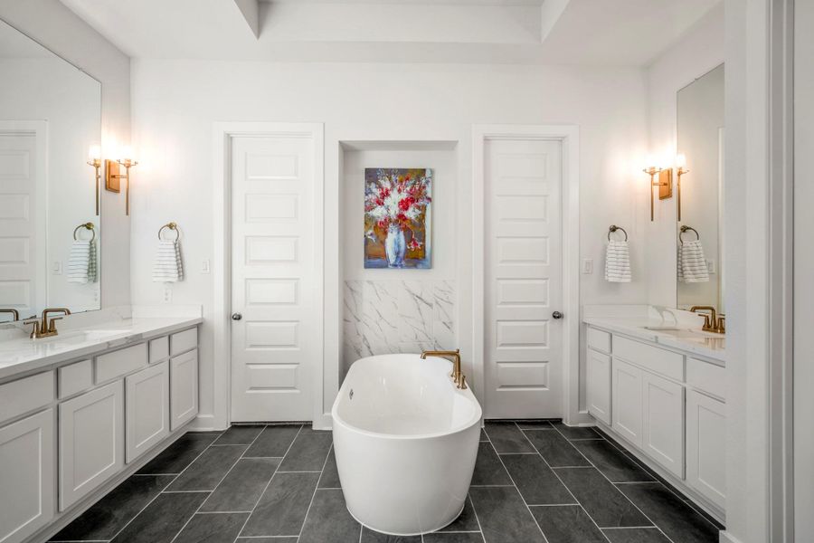 This elegant bathroom features a central freestanding tub, dual vanities with ample counter space, sleek fixtures, and modern lighting. The space is accented with large mirrors and tile flooring, creating a bright and luxurious atmosphere. Enjoy dual walk-in closets in the primary retreat.