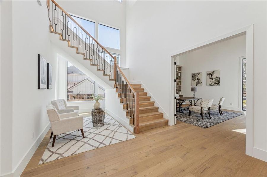 This entryway features a bright and open design, highlighted by large windows that allow for ample natural light. The staircase is a focal point, showcasing light wood steps and a modern railing with intricate black iron detailing. The light wood flooring extends throughout, adding warmth to the space. A wide doorway to the right provides access to an adjacent room, while the high ceilings enhance the spacious and airy atmosphere.