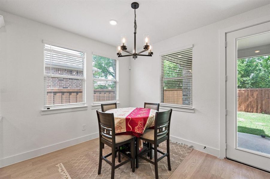 Dining area featuring light wood-finish flooring, a contemporary chandelier, multiple windows with blinds, and a glass-paneled door opening to a private backyard with a wooden fence Dining area featuring light wood-finish flooring, a contemporary chandelier, multiple windows with blinds, and a glass-paneled door opening to a private backyard with a wooden fence