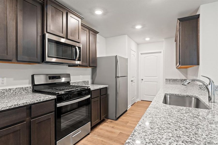 Kitchen with appliances with stainless steel finishes, dark brown cabinets, light stone countertops, light wood-style flooring, and recessed lighting Kitchen with appliances with stainless steel finishes, dark brown cabinets, light stone countertops, light wood-style flooring, and recessed lighting