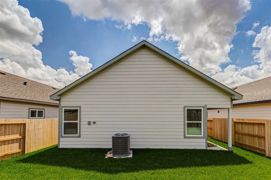 Rear exterior view showcasing the fenced backyard, grassy lawn, exterior windows, HVAC unit, and side-yard access. The private outdoor area provides functional space for outdoor enjoyment with a clean new-construction finish.