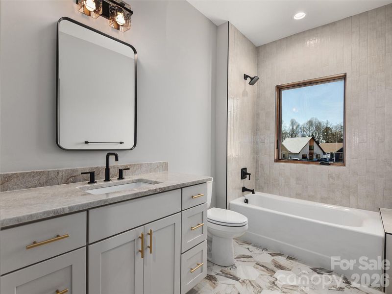 Secondary full bath featuring granite countertop, Wolf cabinetry, designer mirror and lighting, ceramic tile flooring, and tub/shower surround with tiled walls.