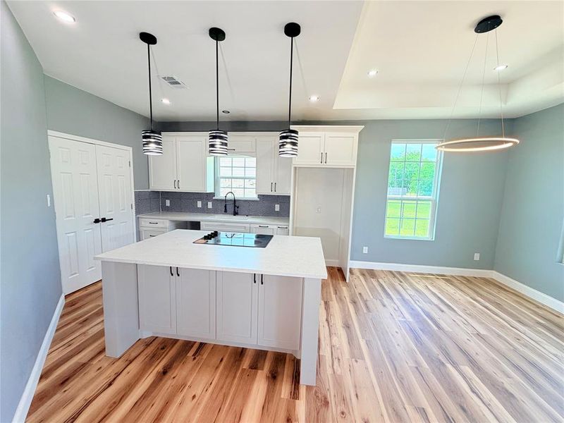 Open-concept kitchen and dining area featuring a kitchen island with built-in cooktop, white shaker cabinetry with dark hardware, a gray subway tile backsplash, and light wood-finish flooring