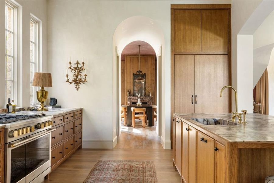Kitchen featuring double oven range, light wood-style floors, a kitchen island with sink, brown cabinets, and light stone countertops