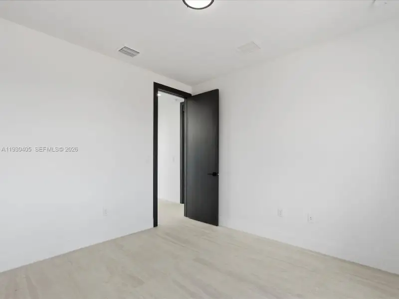 Interior space featuring light-toned wood-finish flooring, a matte black door with matching hardware, and a neutral white color palette Interior space featuring light-toned wood-finish flooring, a matte black door with matching hardware, and a neutral white color palette