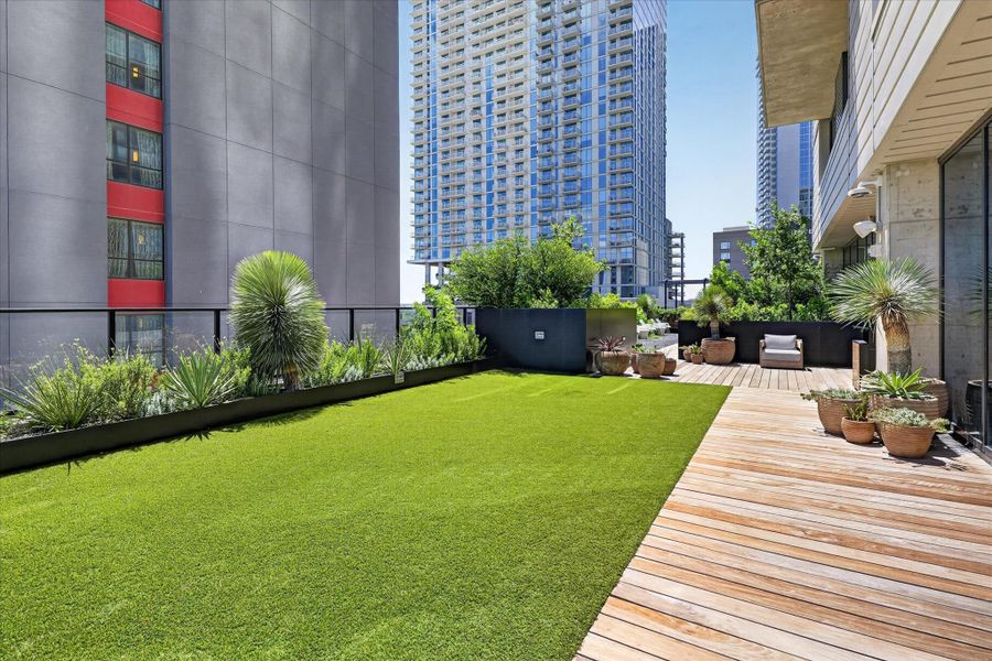 An urban oasis featuring lush artificial turf and warm wooden decking, framed by modern high-rise architecture.