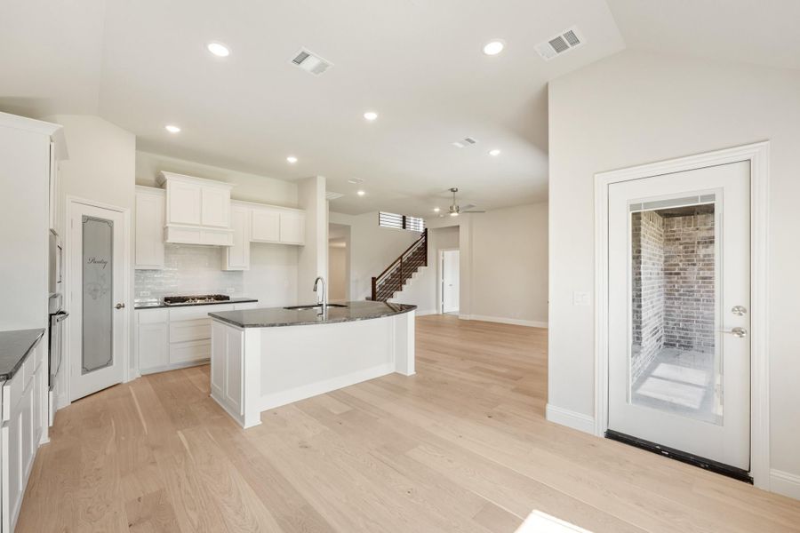 Open kitchen with white cabinets, dark granite island, and light hardwood floors leading to living area and staircase Open kitchen with white cabinets, dark granite island, and light hardwood floors leading to living area and staircase