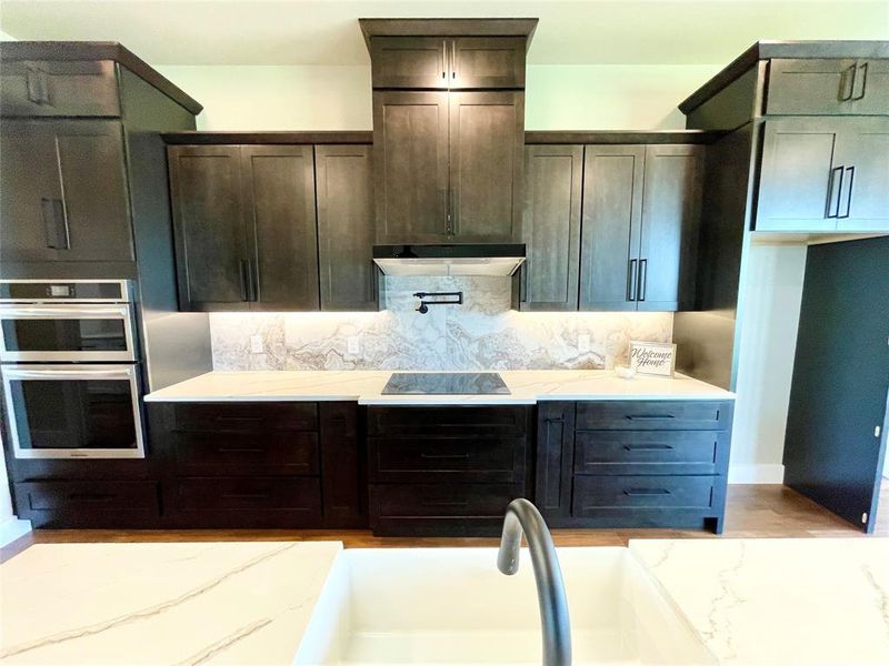 Kitchen with black electric cooktop, dark brown cabinetry, stainless steel double oven, decorative backsplash, and wall chimney range hood