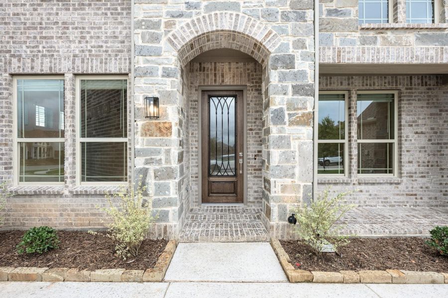 Home entrance with stone arch, wood and glass front door, brick exterior, and small landscaped shrubs on each side Home entrance with stone arch, wood and glass front door, brick exterior, and small landscaped shrubs on each side