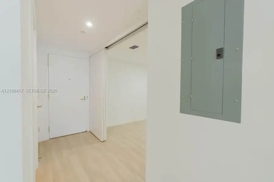 Interior entry featuring light-toned wood-finish flooring, recessed ceiling lighting, a solid-core door with metallic hardware, and an integrated electrical service panel Interior entry featuring light-toned wood-finish flooring, recessed ceiling lighting, a solid-core door with metallic hardware, and an integrated electrical service panel