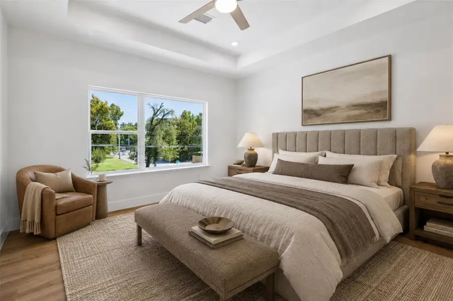 The spacious bedroom offers a bright and peaceful retreat with warm wood-look flooring, crisp neutral walls, and a large window overlooking mature greenery. A tray-style ceiling adds architectural interest, while recessed lighting and a contemporary ceiling fan enhance comfort. The generous dimensions allow room for a complete bedroom suite, additional seating, or a work-from-home area. This photo has been virtually staged for visual reference.