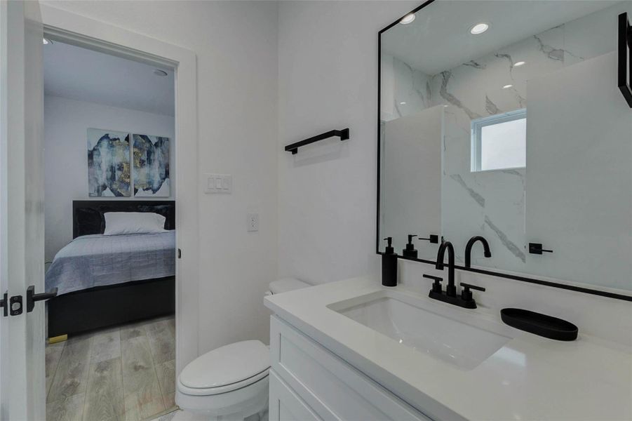 Secondary bath view with white vanity, black faucet, black-framed mirror, modern towel bar, and a clean neutral design. The open layout connects easily to the adjoining bedroom, while the mirror reflects marble-look shower tile for a polished and cohesive look.