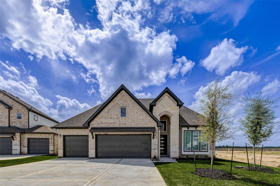 This impressive front elevation showcases a harmonious blend of textures, including light brick and stone accents, complemented by a dramatic arched entryway. The symmetrical design, wide driveway, and three-car garage enhance both convenience and visual appeal. Newly planted trees and fresh landscaping add charm, while the open surroundings provide a sense of space and tranquility, making this home stand out in the neighborhood.