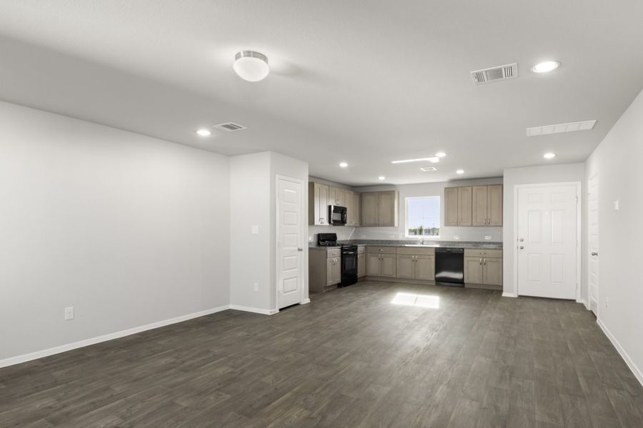 Image of a cottage home living room with dark vinyl flooring, light grey walls, and a L-shaped kitchen in the distance