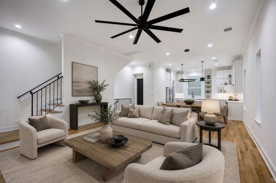 The open main living level offers an impressive sense of space with warm wood flooring, recessed lighting, crown molding, and an oversized contemporary ceiling fan. The modern kitchen sits just beyond, creating an easy connection between cooking, dining, and gathering areas. A sculptural staircase with black iron rails adds architectural character while maintaining the home’s clean, sophisticated style. This photo has been virtually staged for visual reference. The open main living level offers an impressive sense of space with warm wood flooring, recessed lighting, crown molding, and an oversized contemporary ceiling fan. The modern kitchen sits just beyond, creating an easy connection between cooking, dining, and gathering areas. A sculptural staircase with black iron rails adds architectural character while maintaining the home’s clean, sophisticated style. This photo has been virtually staged for visual reference.