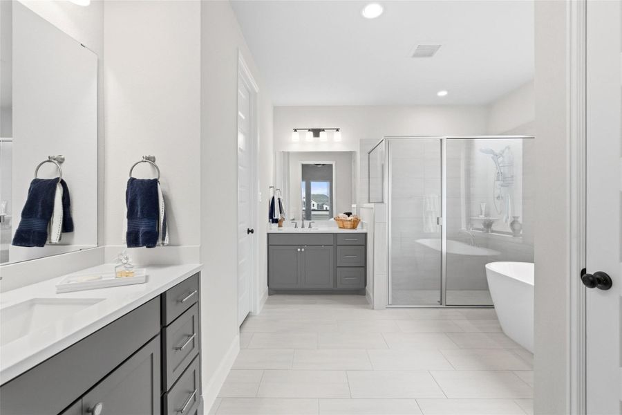 Spacious bathroom featuring light-toned tile flooring, dual vanities with dark cabinetry and light countertops, a glass-enclosed shower, and a freestanding soaking tub