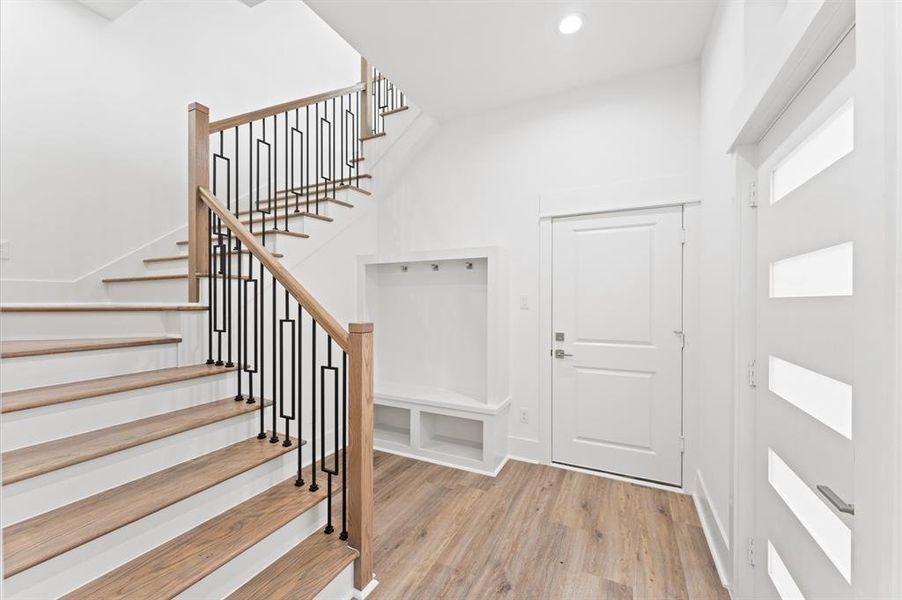 This photo shows a modern entryway with a wooden staircase featuring sleek black metal railing. The space is bright and airy, with light wood flooring and white walls. There's a built-in bench with hooks for storage and a white door with horizontal glass panels.