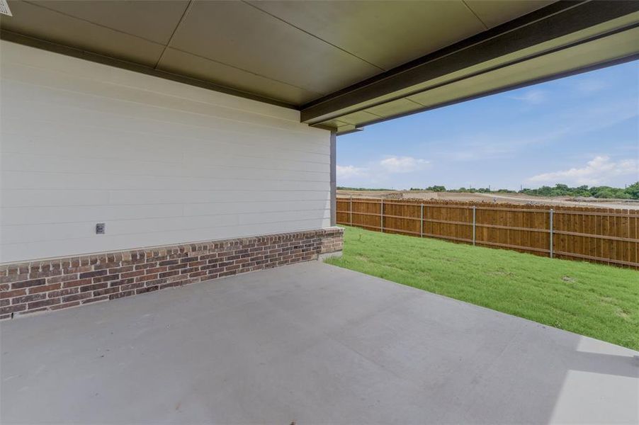 Covered concrete patio with white siding and brick veneer, bordered by a wooden privacy fence and a grass lawn Covered concrete patio with white siding and brick veneer, bordered by a wooden privacy fence and a grass lawn