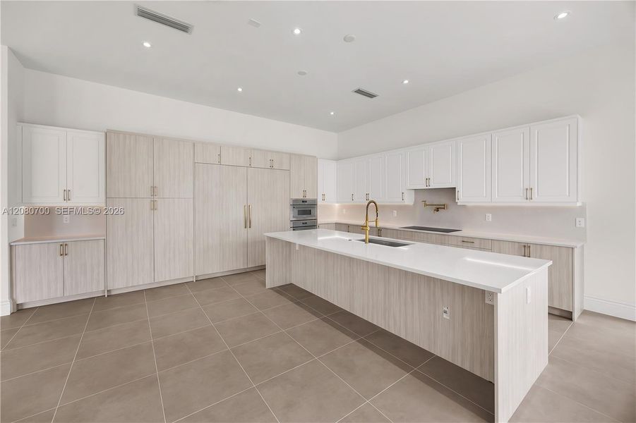 Expansive kitchen featuring a large central island with a sink, extensive white cabinetry, light wood-finish cabinetry, built-in stainless steel appliances, and sleek tile flooring