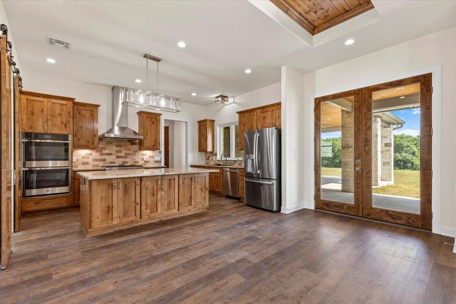 Kitchen featuring stainless steel appliances, wall chimney exhaust hood, brown cabinetry, a kitchen island, and decorative backsplash