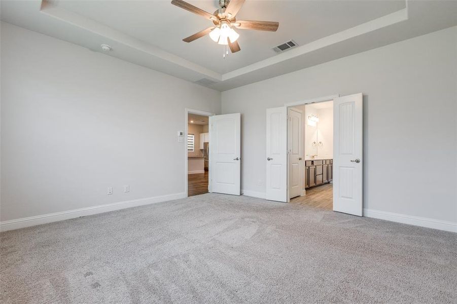 Unfurnished bedroom with light carpet, a tray ceiling, baseboards, a ceiling fan, and ensuite bathroom