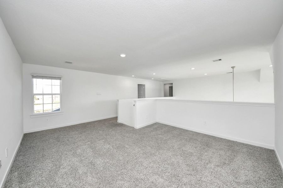 A spacious, carpeted loft area with recessed lighting. It features a window for natural light and an open railing overlooking the space below, offering a versatile area for various uses.