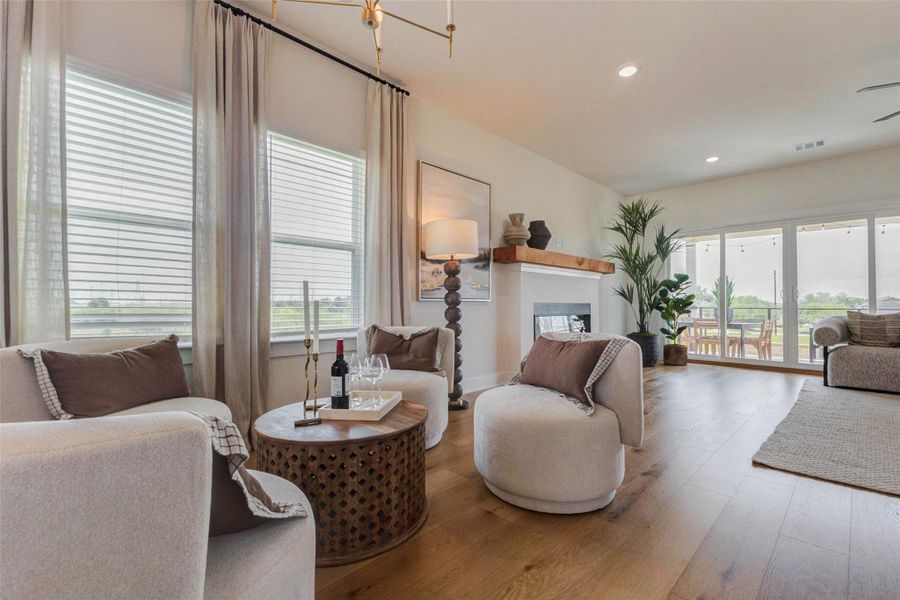 This inviting living space offers a perfect balance of comfort and style, featuring warm wood flooring, soft neutral tones, and abundant natural light. A cozy seating arrangement creates an intimate gathering area, while the fireplace adds a focal point with a touch of warmth and charm. Large sliding doors open to the outdoor space, enhancing the home’s indoor-outdoor living experience. The picture shown is from the model home with the same floor plan, Rev floor plan.