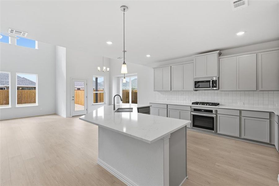 Kitchen featuring gray cabinetry, stainless steel appliances, a kitchen island with sink, light wood-type flooring, and light stone countertops
