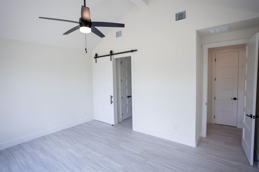 Unfurnished bedroom with a barn door, high vaulted ceiling, light wood-style flooring, a ceiling fan, and beamed ceiling Unfurnished bedroom with a barn door, high vaulted ceiling, light wood-style flooring, a ceiling fan, and beamed ceiling