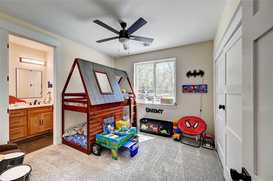 This bright children's room features a whimsical loft bed, playful decor, and a cozy carpeted floor. It includes a ceiling fan and ample natural light from a large window. A convenient en-suite bathroom with modern fixtures is also accessible.