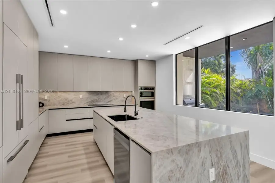 Kitchen featuring flat-panel cabinetry, a waterfall-edge island with a stone countertop, an undermount sink with a gooseneck faucet, built-in wall ovens, and floor-to-ceiling windows Kitchen featuring flat-panel cabinetry, a waterfall-edge island with a stone countertop, an undermount sink with a gooseneck faucet, built-in wall ovens, and floor-to-ceiling windows
