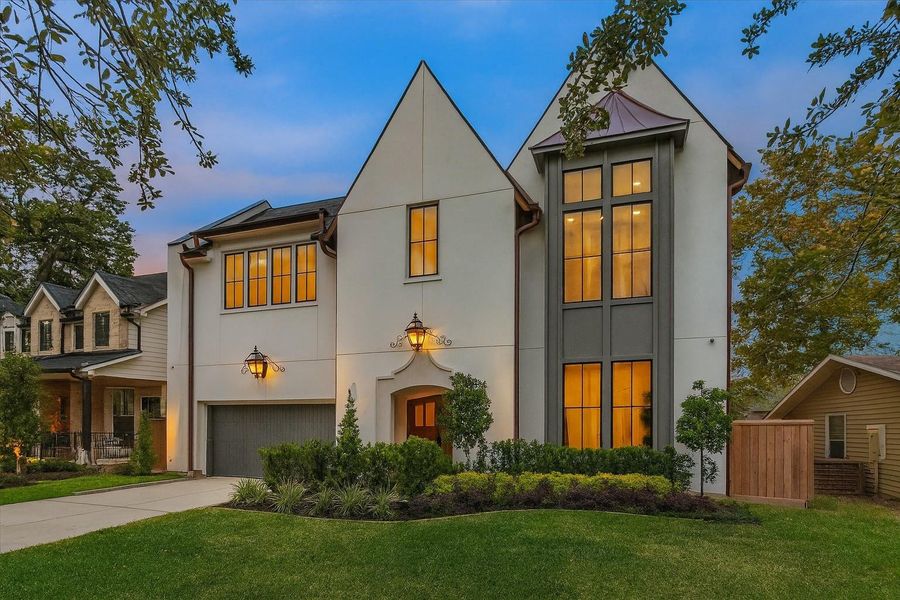 The front elevation introduces the home’s refined architectural style with its dramatic twin gables, smooth stucco finish, rounded downspouts, and floor-to-ceiling divided-light windows that bathe the interior in natural light. A custom stone-trimmed arched entry and warm exterior lantern create an inviting focal point, while professional landscaping enhances the modern European profile. The front elevation introduces the home’s refined architectural style with its dramatic twin gables, smooth stucco finish, rounded downspouts, and floor-to-ceiling divided-light windows that bathe the interior in natural light. A custom stone-trimmed arched entry and warm exterior lantern create an inviting focal point, while professional landscaping enhances the modern European profile.
