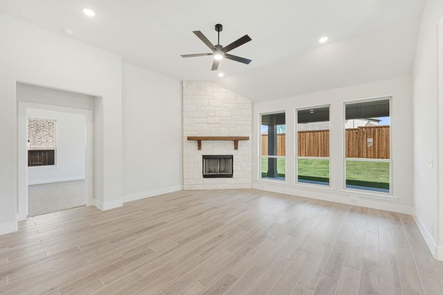 Open living room with stone fireplace, ceiling fan, light wood floors, and large backyard-facing windows