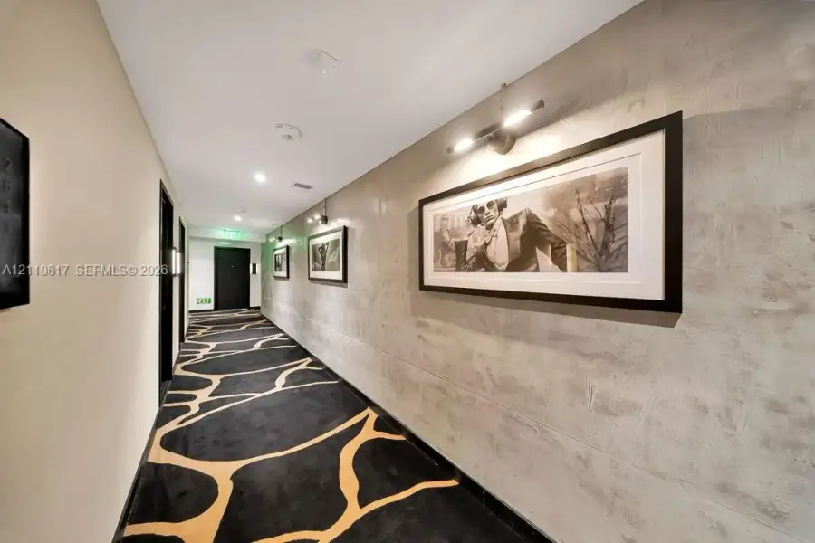 Hallway featuring textured grey wall surfaces, patterned carpeting, and directional wall-mounted lighting Hallway featuring textured grey wall surfaces, patterned carpeting, and directional wall-mounted lighting