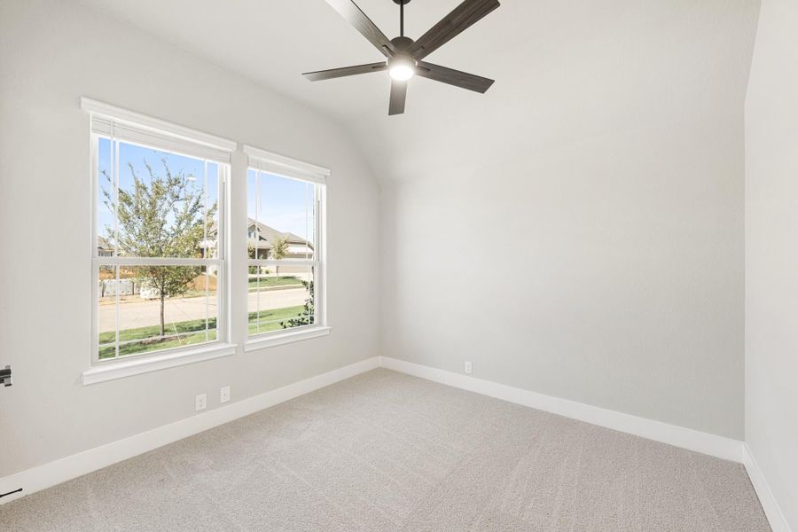 Empty bedroom with vaulted ceiling, ceiling fan, carpet flooring, and two windows overlooking a neighborhood street.