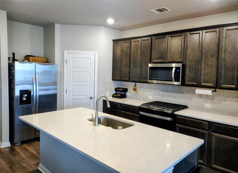 Kitchen with dark brown cabinets, appliances with stainless steel finishes, light stone countertops, a center island with sink, and recessed lighting