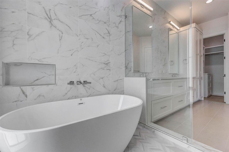 Striking freestanding soaking tub set against marble-look tile with a built-in niche and wall-mounted fixtures. Herringbone-pattern tile flooring adds subtle texture and visual interest, delivering a refined spa-like focal point within the primary bath.
