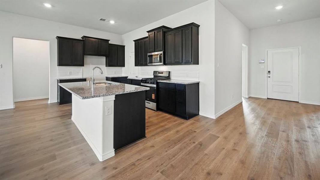 Kitchen featuring dark cabinets, stainless steel appliances, light stone counters, a center island with sink, and decorative backsplash