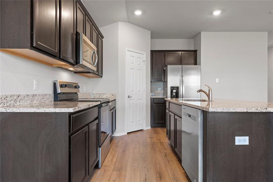 Kitchen featuring appliances with stainless steel finishes, dark brown cabinets, light wood-style flooring, light stone counters, and a kitchen island with sink Kitchen featuring appliances with stainless steel finishes, dark brown cabinets, light wood-style flooring, light stone counters, and a kitchen island with sink