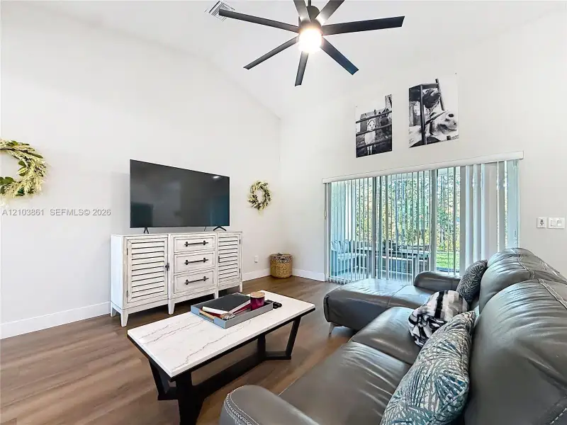 Vaulted ceiling featuring a central matte black ceiling fan, wood-finish flooring, and sliding glass patio doors with vertical blinds