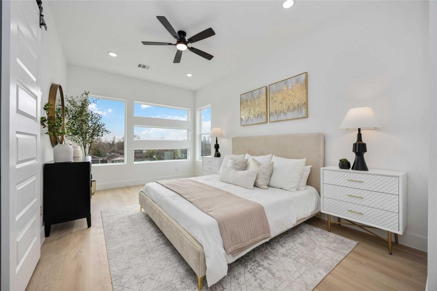 This beautifully appointed bedroom showcases expansive picture windows that bathe the space in natural light, complemented by warm wood-look flooring, modern recessed lighting, and a sleek ceiling fan; the room is elegantly styled with contemporary furnishings that highlight its spacious and serene ambiance.