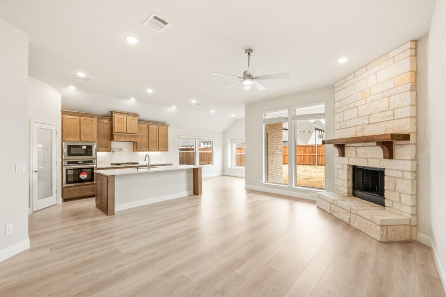 Open-concept living area with stone fireplace, kitchen island, wood cabinets, and light hardwood floors Open-concept living area with stone fireplace, kitchen island, wood cabinets, and light hardwood floors