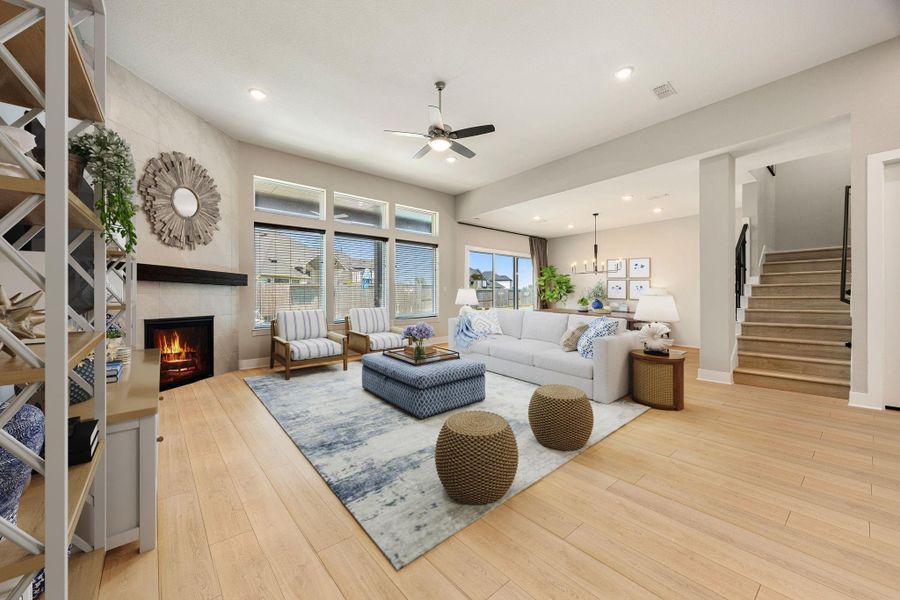 This inviting living area showcases warm wood-look flooring, a cozy corner fireplace with a modern tile surround, and a wall of windows that floods the space with natural light while offering views of the backyard. The open-concept layout flows seamlessly into the dining area and kitchen, while recessed lighting and neutral tones create a bright, welcoming atmosphere. A staircase with sleek black railing adds a touch of sophistication and leads to the upper level.