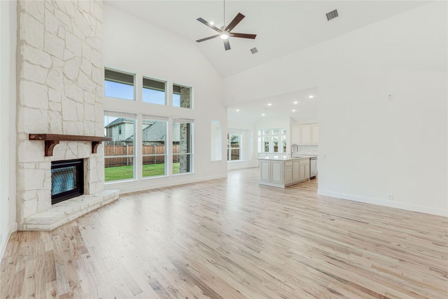 Open living room with floor-to-ceiling stone fireplace, hardwood floors, high ceiling, and open kitchen in background